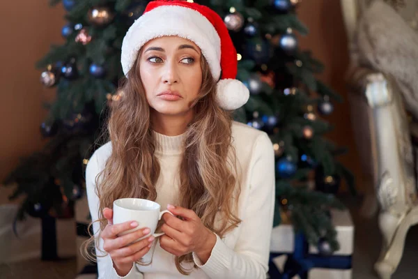 Christmas Woman dressed white sweater Santa hat sitting floor near christmas tree  Caucasian female relaxing winter holiday home interior drink hot morning coffee and upset. It was a bad year Boring