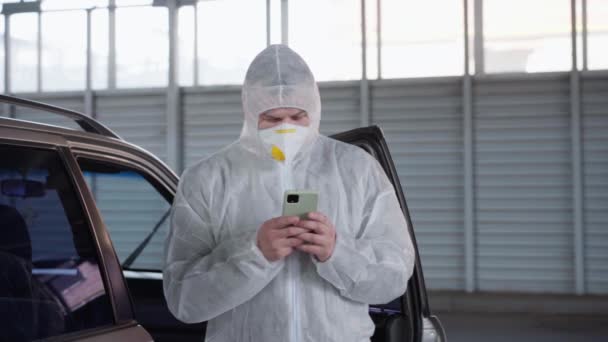 Man arts gekleed witte beschermende overalls in de buurt van auto rusten met behulp van de smartphone — Stockvideo