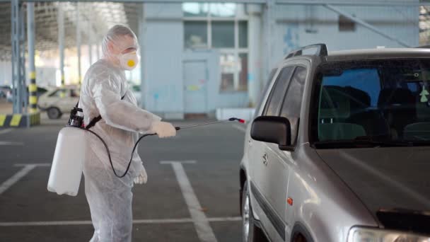 Man dressed white protective overalls spraying vehicle antibacterial sanitizer sprayer on quarantine Caucasian person protective suit disinfects car on parking during covid pandemic — Stock Video