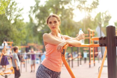 Sportif bir kadın yaz günü spor sahasında durup bir şişe orta boy su ile antrenman yaptıktan sonra dinleniyor. Kafkasyalı atletik kadın antrenmandan sonra biraz dinlenir.