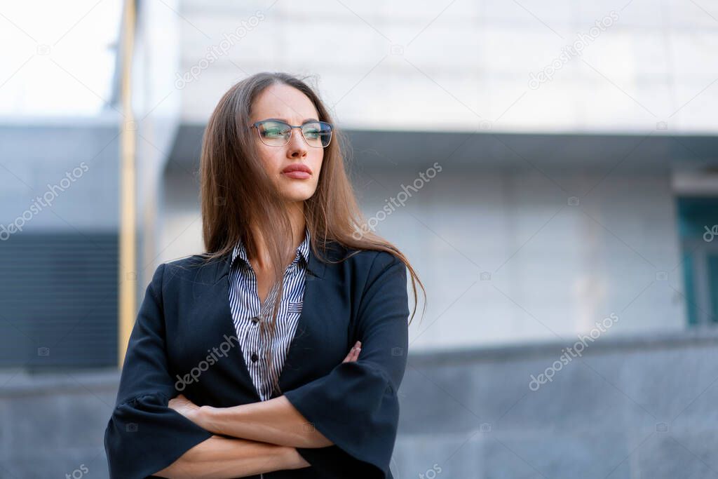 Business woman with long hair dressed black jacket eyeglasses standing outdoor near corporate office building hands folded Beautiful caucasian female business person portrait on city street Side view