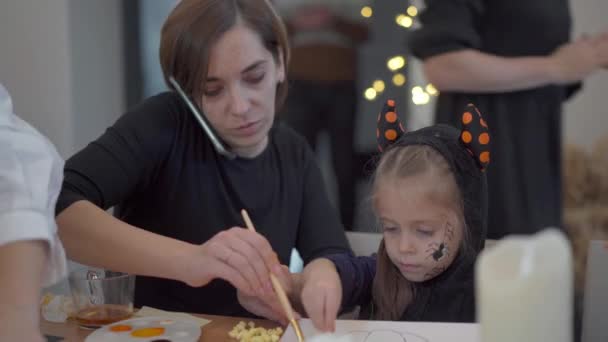 Malá dívka oblečená Halloween karneval kostým s její matkou sedí na stole kreslení ručně Halloween dekorace na party — Stock video