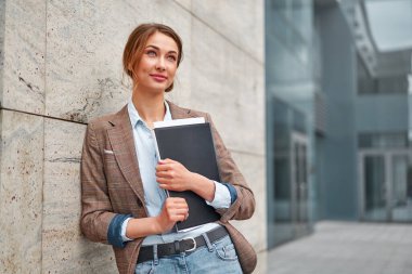 Elinde bir dosya dolusu belgeyle sarışın bir iş kadını rüya görüyor ve kafasını kaldırıyor. İş konsepti. Arka planda bulanık bir iş merkezi görüntüsü var..