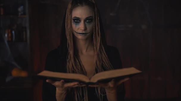 Концепция Хэллоуина. Ведьма в черном капюшоне с дредами, стоящими в темном подземелье, использует магическую книгу для заклинаний, дующих волшебную пыль в камеру. — стоковое видео