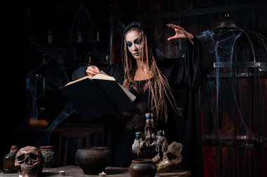 Cadılar Bayramı konsepti Kara başlıklı cadı karanlık zindan odasında büyü yapmak için sihirli kitap kullanıyor. Dişi büyücü büyücü Gotik iç iskelet, kafes, örümcek ağı