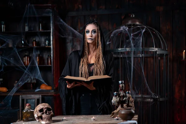 ハロウィンのコンセプト 魔女が黒いフードにドレッドロックが立っているダークダンジョンルーム呪いを引き起こすための魔法の本を保持します 女性のネクロマンサーウィザード頭蓋骨とカメラを探して ケージ クモの巣テーブル — ストック写真