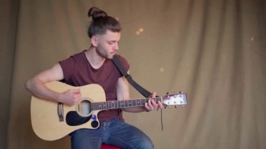 Akustik gitar çalan yakışıklı genç adam. Oturma odası kumaş arka planda.