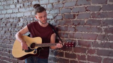 Akustik gitarlı bir adam tuğla duvara yaslanmış müzik çalıyor, hayatın tadını çıkarıyor.