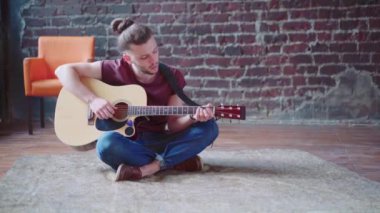 Akustik gitar çalan yakışıklı genç adam oturma odasında oturuyor.