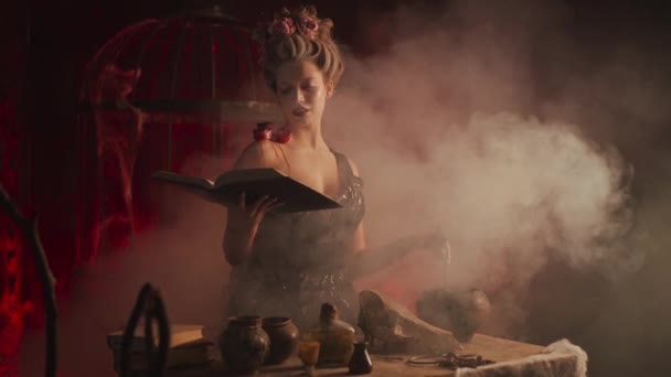 Halloween Hexe mit Hexenkessel und Zauberbuch. Schöne junge Frau zaubert, macht Hexerei — Stockvideo