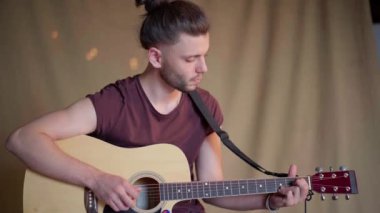 Akustik gitar çalan yakışıklı genç adam. Oturma odası kumaş arka planda.