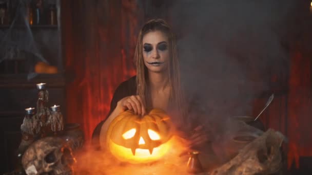 Halloween concept. Black witch holding Halloween pumpkin with carved smily face in hand sitting on the table dark room — Stockvideo