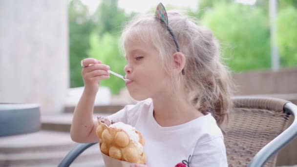 Liten flicka äter bubbla våffla med glass sitter utomhus café sommardag — Stockvideo