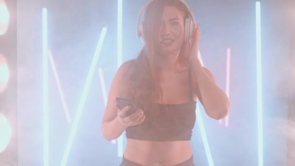 Artistic young woman take selfie on smartphone headphones enjoy music dancing in backlit fog smoke indoors. Confident graceful Caucasian female dancer — стокове відео