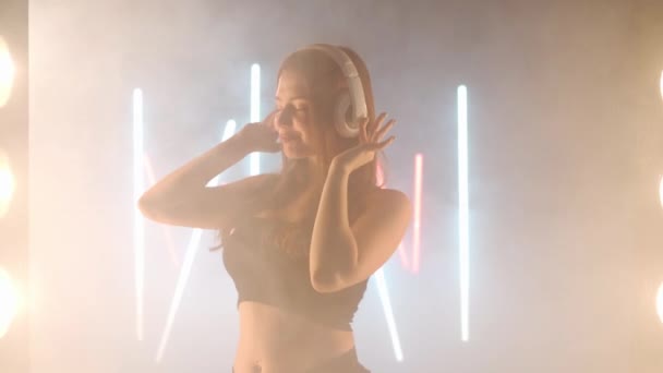Художня молода жінка - навушники, що танцюють у засвіченому тумані. Впевнена, що кавказька танцівниця репетирує сучасну виставу з балетними рухами. — стокове відео