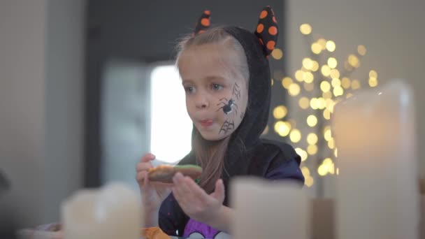 Klein meisje eet glazuur handgemaakte Halloween koekjes gekleed Halloween carnaval kostuum met hoorns — Stockvideo