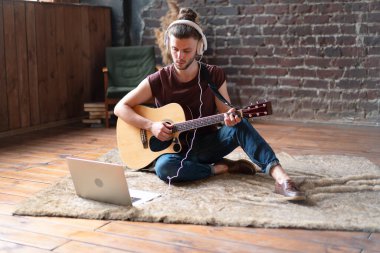 Kafkasyalı genç yetişkin uzak öğretmen bilgisayarlı mobil cihazıyla online gitar çalmayı öğreniyor. Yakışıklı hippi dizüstü bilgisayarla müzik öğretiyor, uzaktan eğitim kavramı 