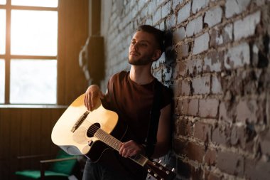 Akustik gitarı olan, tuğla duvara yaslanmış, müzik çalan, hayatın tadını çıkaran yakışıklı beyaz gitarist ev yapımı, müzikal enstrüman çalıyor. 