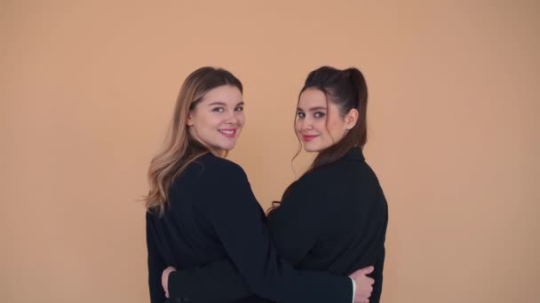 Two young business women stand in the studio backs to camera hugging turns around and blows kiss then laughs merrily. Business partners concept. — Stock Video