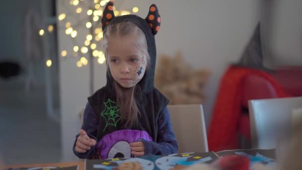 Маленька дівчинка прикрашає глазур'ю печиво ручної роботи на Хеллоуїн, одягнене в карнавальний костюм на Хелловін з рогами — стокове відео