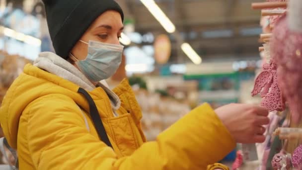 Sarı kışlık ceketli bir kadın alışveriş merkezinde Noel süslerini seçiyor. Tıbbi koruyucu maskeli yetişkin beyaz kadın yeni yıl süsü alıyor. — Stok video