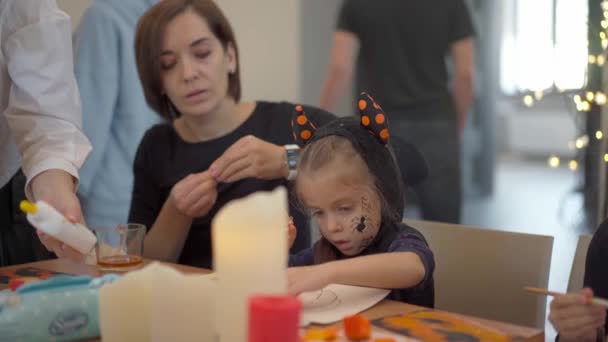 Cadılar Bayramı karnavalı kostümü giymiş küçük bir kız. Annesi masada oturmuş, Cadılar Bayramı süslemelerini çiziyor. — Stok video