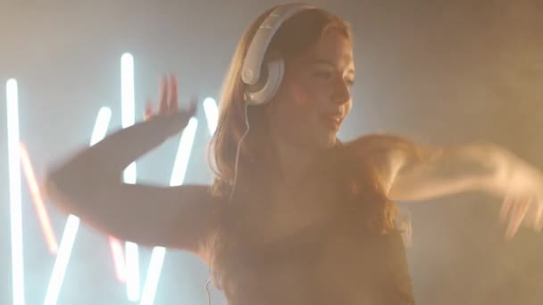 Artistic young woman headphones dancing in backlit fog smoke indoors. Confident graceful Caucasian female dancer rehearsing modern performance with ballet movements — Stock video