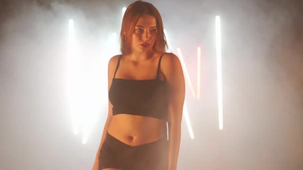 Mujer joven artística bailando en humo de niebla retroiluminada en interiores. Bailarina femenina caucásica confiada ensayando actuación moderna con movimientos de ballet — Vídeo de stock