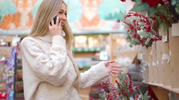 Shopping de Noël, femme européenne choisir des branches pour la couronne de Noël à l'intérieur dans le supermarché parler au téléphone. Femme caucasienne achetant décor de Noël sur le marché de vacances — Video