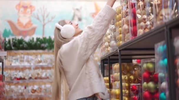 Karácsonyi vásárlás. A nő a karácsonyi dekorációt választja a szupermarketben. Fehér nő szőrme fejhallgatóban, fehér pulóverben, karácsonyi díszt vesz. Készülj a téli szünetre! — Stock videók