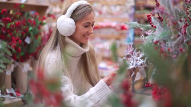 Compras de Navidad, mujer europea elegir ramas para la corona de Navidad interior en el supermercado. Mujer caucásica compra decoración de Navidad en el mercado de vacaciones, auriculares de piel vestida y suéter blanco. — Vídeos de Stock