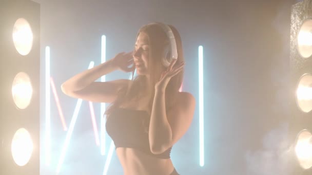 Auriculares de mujer joven artística bailando en humo de niebla retroiluminada en interiores. Bailarina femenina caucásica confiada ensayando actuación moderna con movimientos de ballet — Vídeos de Stock