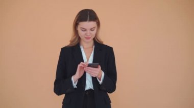 Genç iş kadını, sarı saçlı, siyah takım elbiseli, cep telefonu tutan, gülen, bej arka planda izole edilmiş bir stüdyo.