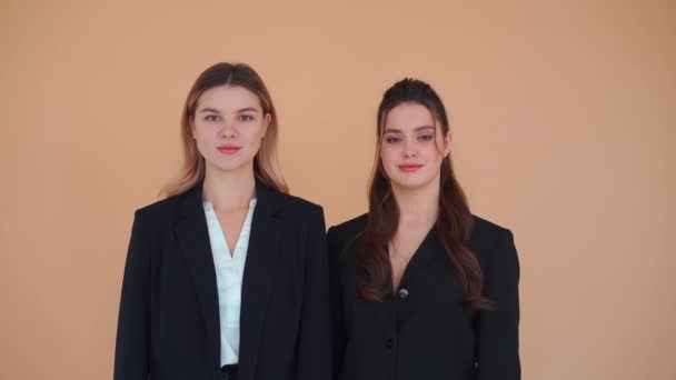 Dos jóvenes mujeres de negocios vestidas de negro traje formal de pie estudio de fondo beige mirando cámara. Concepto de asociación empresarial — Vídeo de stock