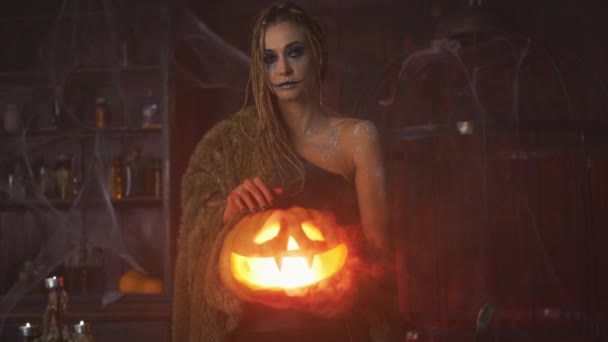 Концепция Хэллоуина. Черная ведьма держит тыкву на Хэллоуин с вырезанным улыбающимся лицом в руке, стоящим в темной комнате — стоковое видео