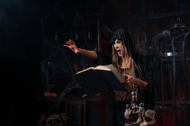 Cadılar Bayramı konsepti Karanlık zindan odasında duran siyah başlıklı cadı büyü yapmak için sihirli kitap kullanıyor. Büyücü kadın büyücü Gotik iç mekan elini öne doğru attı.