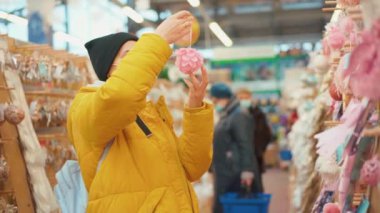Sarı kışlık ceketli bir kadın alışveriş merkezinde Noel süslerini seçiyor. Tıbbi koruyucu maskeli yetişkin beyaz kadın yeni yıl süsü alıyor. 