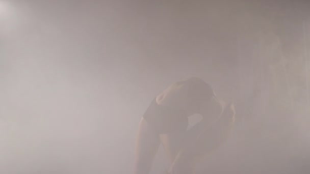 Mujer joven artística bailando en humo de niebla retroiluminada en interiores. Bailarina femenina caucásica confiada ensayando actuación moderna con movimientos de ballet — Vídeos de Stock