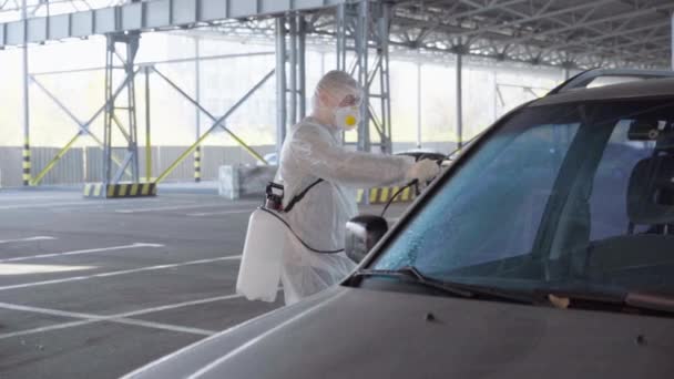 Hombre vestido blanco mono protector pulverización vehículo antibacteriano desinfectante pulverizador en cuarentena traje protector persona caucásica desinfecta coche en estacionamiento durante pandemia covid — Vídeo de stock