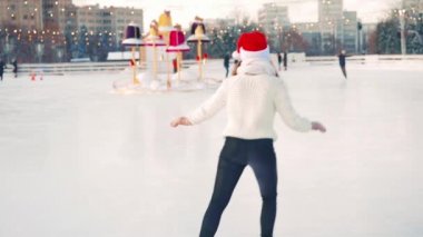 Gülümseyen genç kadın Noel Baba, buz pateni pistinde beyaz kazak giyerek buz pateni yapıyor. Noel tatili, aktif kış tatili el bilgisayarı etkisi. Amatör beyaz kadın buz patencisi. 