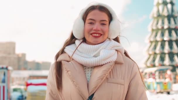 Mladá žena oblečená kožešinové ucho a bílá pletená šála stojící venkovní zimní sezóna na ulici města s vánoční stromeček na pozadí — Stock video