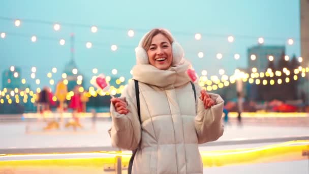 Frau mit Ohrenschützern steht am Winterferienabend im Freien in der Nähe der Eisbahn am zentralen Stadtplatz. Menschen, die im Hintergrund mit Weihnachtslicht Schlittschuh laufen. — Stockvideo