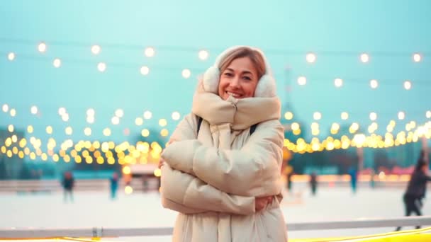 Frau gefroren auf winterlicher Straße umarmt sich in warmer Winterkleidung und Ohrenschützer, die draußen auf der Eisbahn der Stadt stehen und Weihnachtsbeleuchtung auf dem Hintergrund — Stockvideo