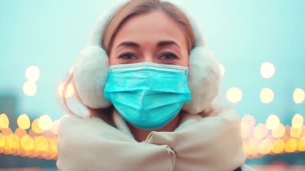 Mujer optimista de confianza usar máscara facial de protección médica durante covid 19 pandemia de pie fuera de la calle de la ciudad de invierno con iluminación de Navidad en el fondo mirando cámara. — Vídeo de stock