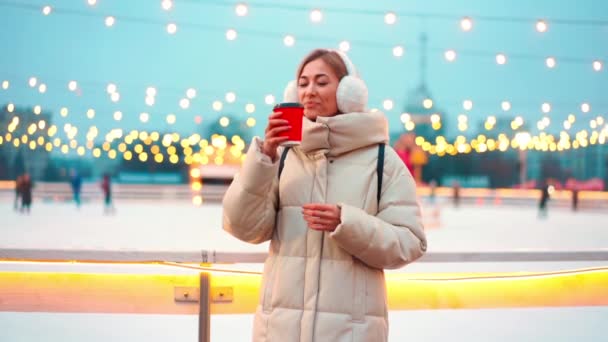 Mujer adulta beber café para ir en gorra de papel de pie fuera en la calle de la ciudad pista de hielo y fondo claro de Navidad. Vacaciones de invierno hermosa noche en el centro de Invierno. Ocio de invierno. — Vídeos de Stock