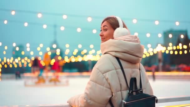 Winterzeit. Heiligabend steht eine Frau vor der Eisbahn und blickt zurück und lächelt. Weihnachtsbeleuchtung und Dekoration Hintergrund. Winterferienzeit — Stockvideo