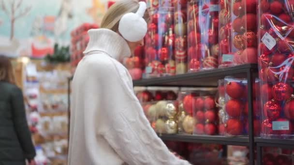 Kerstinkopen. Vrouw kiest voor kerstdecor in supermarkt. Blanke vrouw in bont koptelefoon gekleed witte trui kopen kerstversiering. Bereid je voor op de winter Vakantie — Stockvideo