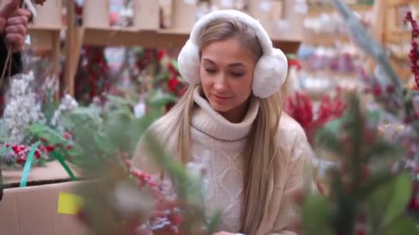 Compras de Navidad, mujer europea elegir ramas para la corona de Navidad interior en el supermercado. Mujer caucásica compra decoración de Navidad en el mercado de vacaciones, auriculares de piel vestida y suéter blanco. — Vídeos de Stock