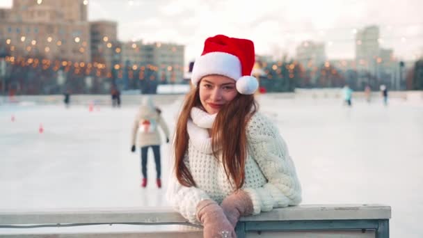 Gülümseyen genç kadın Noel Baba, buz pateni pistinde beyaz kazak giyerek buz pateni yapıyor. Noel tatili, aktif kış tatili el bilgisayarı etkisi. Amatör beyaz kadın buz patencisi. — Stok video