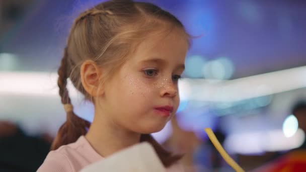 Κοριτσάκι με κοτσιδάκια να τρώει τηγανητές πατάτες. Κοντινό πλάνο πορτρέτο καυκάσιος θηλυκό 5 χρόνια τρώνε fast food σε ένα εστιατόριο το βράδυ με ελαφριά μουσική στο παρασκήνιο — Αρχείο Βίντεο
